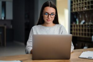 Brunette focused young woman in eyeglasses working with laptop at home